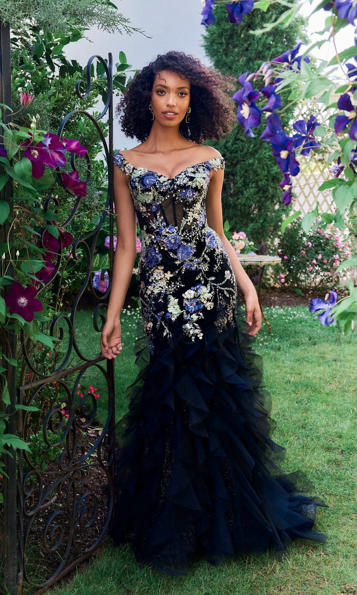 A woman in the Morilee 49148 long prom dress with off-shoulder floral embroidery stands by an ornate garden gate, surrounded by purple flowers and lush greenery.
